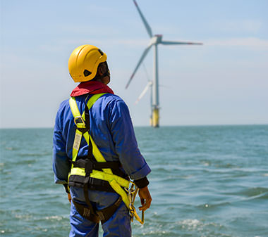 a worker in helmet and harness looking out at a wind turbine on the water